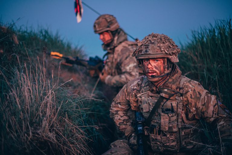 Two military soldiers in camouflage uniforms and helmets sit in tall grass at dusk, one holding a rifle and the other looking ahead with a serious expression, reflecting the challenges faced by veterans during resettlement.