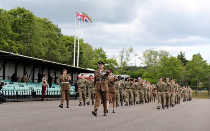 A group of uniformed soldiers, including a Pathfinder veteran, march in formation past empty green seating under a cloudy sky, with two flags flying in the background.