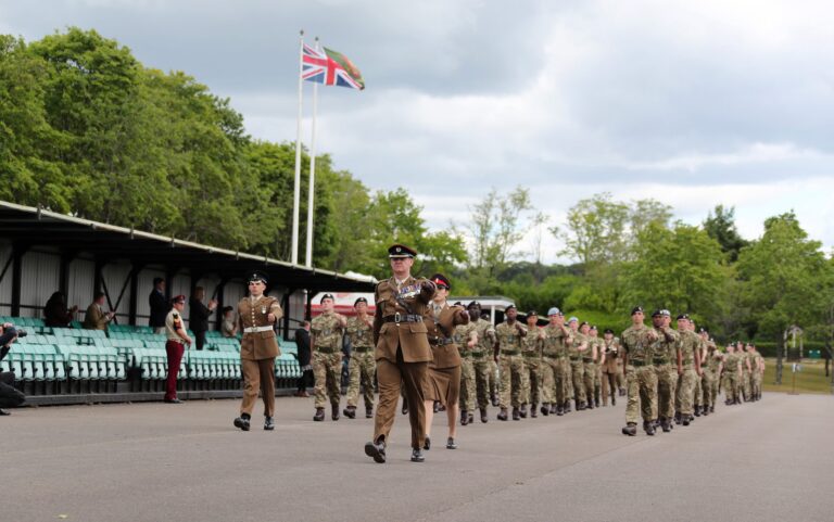 A group of uniformed soldiers, including a Pathfinder veteran, march in formation past empty green seating under a cloudy sky, with two flags flying in the background.