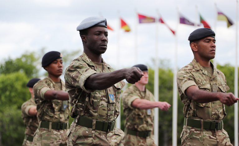 A group of uniformed soldiers, including Royal Navy personnel, marches in formation outdoors with flags in the background.