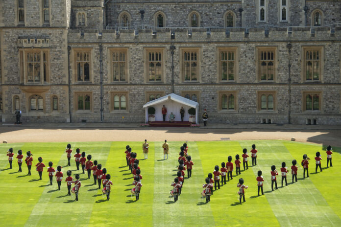 British military guards in red uniforms and bearskin hats perform a formation on a lawn before a stone building with a tented platform and two people standing under it.