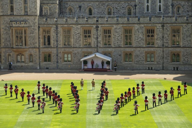 British military guards in red uniforms and bearskin hats perform a formation on a lawn before a stone building with a tented platform and two people standing under it.