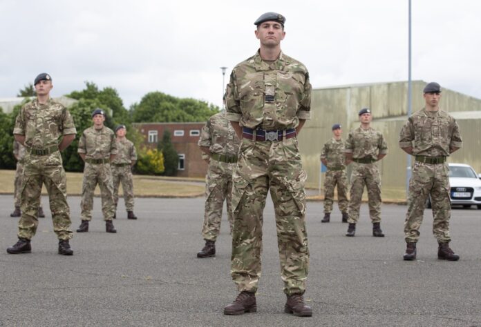 A group of Army soldiers in camouflage uniforms stands at attention outdoors on a paved area, with buildings and trees in the background.