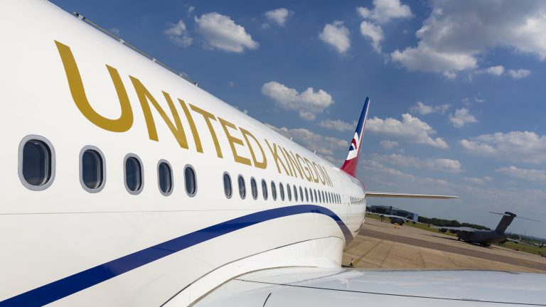 A large military aircraft with "UNITED KINGDOM" on its side is parked on an airport tarmac under a partly cloudy sky, ready to support resettlement efforts for Pathfinder missions.