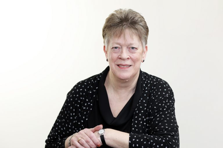 An older woman with short gray hair, a Royal Navy veteran, sits smiling with her arms crossed on a table in front of a plain white background, wearing a black top and patterned cardigan, reflecting on her resettlement journey.