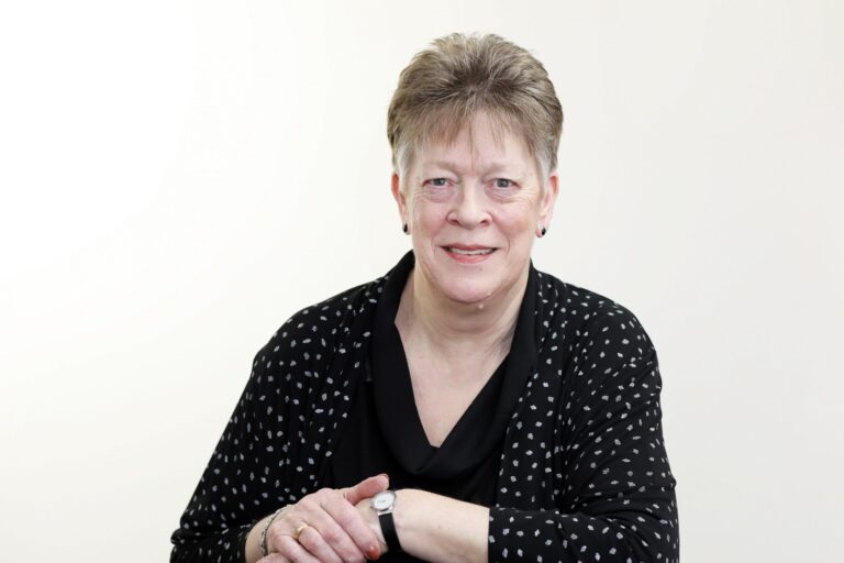 An older woman with short gray hair, a Royal Navy veteran, sits smiling with her arms crossed on a table in front of a plain white background, wearing a black top and patterned cardigan, reflecting on her resettlement journey.