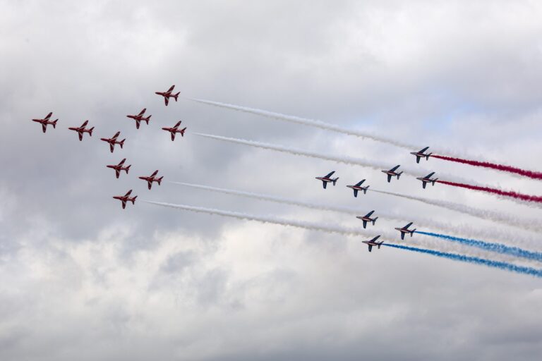 Two aerobatic teams from the Army and Royal Navy fly in formation, leaving trails of white, red, and blue smoke against a cloudy sky.