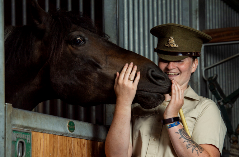 A person in a military uniform stands by a dark horse in a stable, smiling as they gently hold the horse's face with both hands—embodying the caring spirit of military resettlement and new pathfinder journeys.
