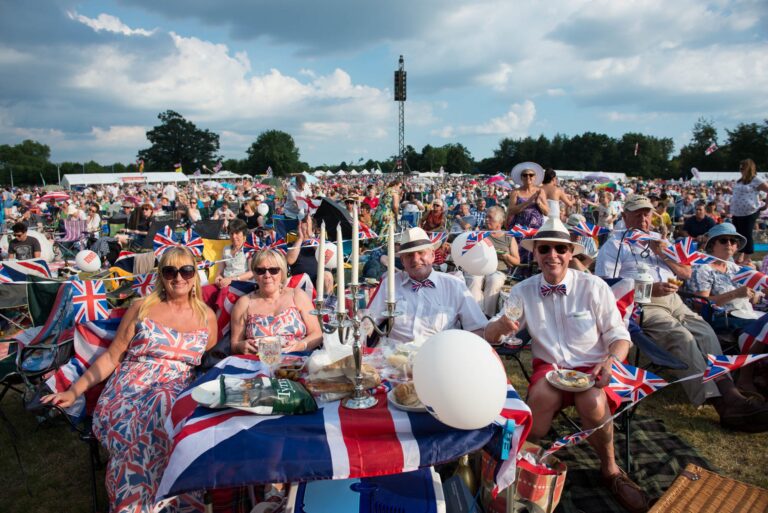 A large outdoor crowd with people in summer clothes sits at a picnic table decorated with Union Jack flags, enjoying food at a British festival celebrating veterans and honoring the Royal Navy.