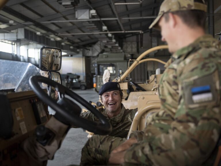 Two soldiers in camouflage uniforms interact near a military vehicle inside a garage; one, a Pathfinder, is sitting in the driver’s seat, smiling, while the other stands beside him.