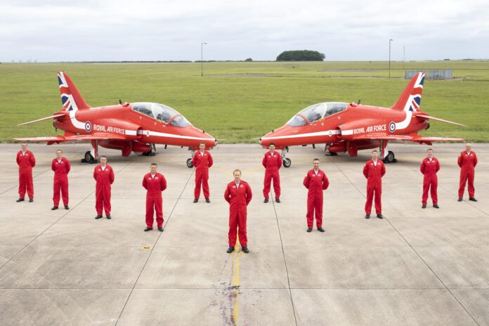 A group of eleven people in red flight suits stand spaced apart on a tarmac in front of two red Royal Air Force jets, with grassy fields in the background—showcasing military precision and teamwork.