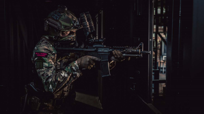 A soldier in camouflage uniform and helmet, wearing tactical gear and night vision goggles, aims a rifle inside a dimly lit building—a true Pathfinder showcasing the skills of a Veteran.