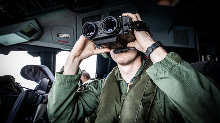 A person in a military uniform uses binoculars inside an Army aircraft cockpit.