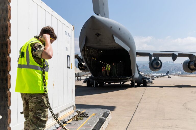 A person in Army uniform and high-visibility vest stands next to a shipping container near a large cargo aircraft with its rear ramp open on an airport tarmac.