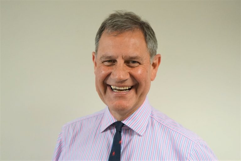 Smiling middle-aged Army veteran, wearing a striped shirt and dark tie, stands against a plain light background.