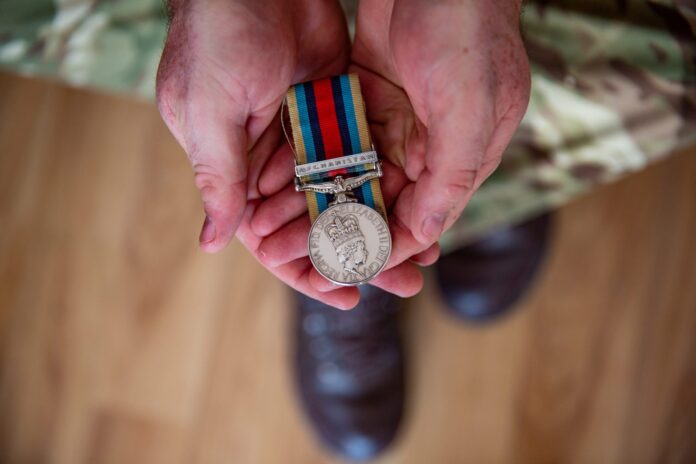A person in camouflage uniform holds a military medal with a striped ribbon in their hands, viewed from above—an emblem of honor marking their service and resettlement beyond the Military or Royal Navy.