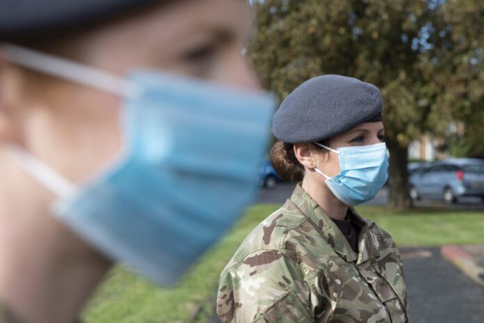 Two military personnel in camouflage uniforms and grey berets stand outdoors, both wearing blue surgical face masks, possibly discussing resettlement options for veterans.