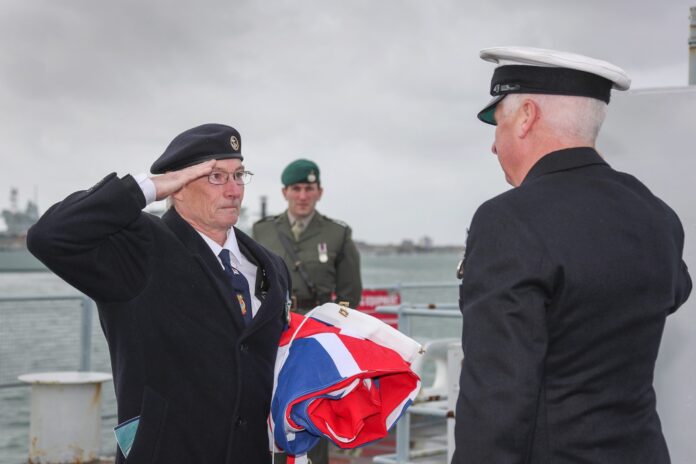 A Royal Navy veteran salutes another officer while holding a folded flag, with a third uniformed person standing in the background on a ship.