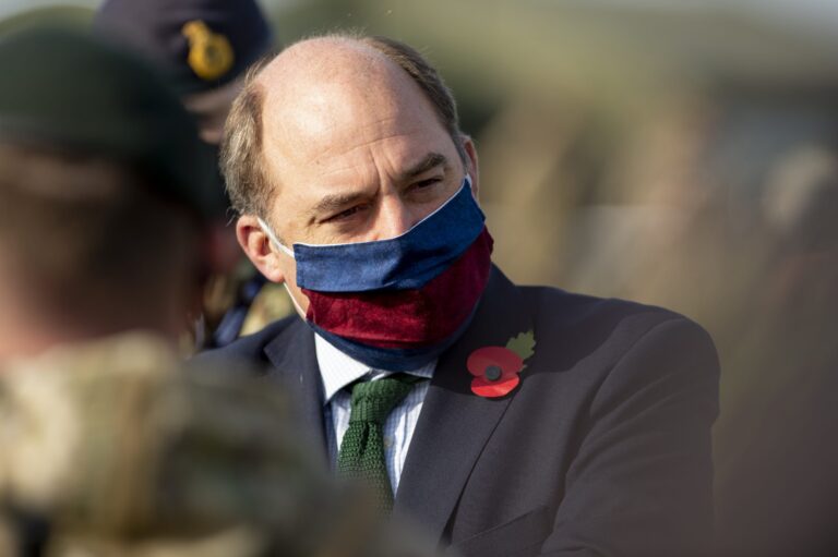 A man wearing a red and blue face mask, suit, and poppy pin stands outdoors among people in Royal Navy uniforms.