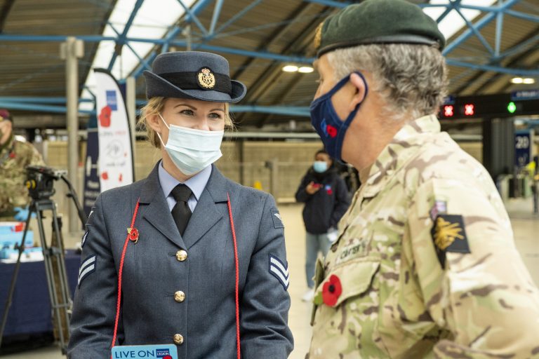 A veteran in an air force uniform and a man in army camouflage, both wearing masks and poppies, talk inside a train station.