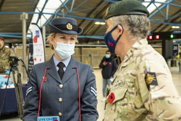 A veteran in an air force uniform and a man in army camouflage, both wearing masks and poppies, talk inside a train station.