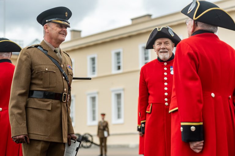 A military officer in uniform discusses resettlement with two men in red coats and black hats during an outdoor event at a historical building.