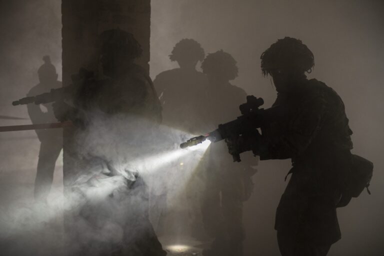 Four military soldiers with rifles and tactical gear move cautiously through a smoky, dimly lit area, with one veteran using a flashlight mounted on their weapon.