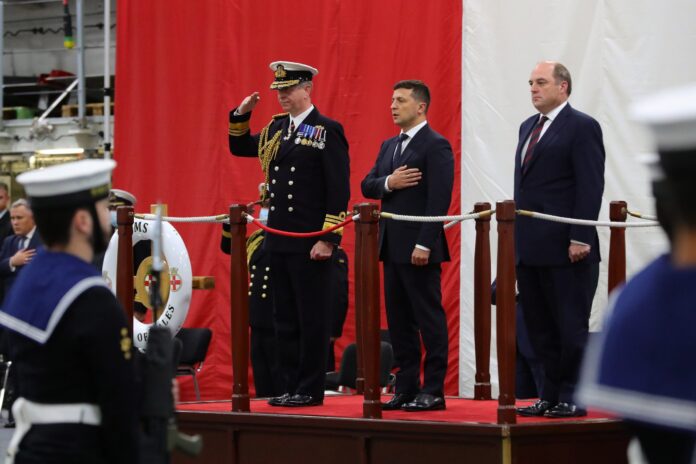 Three men in formal attire stand on a platform; one salutes in a military manner, one holds a hand to his chest, and one stands with arms at his sides. Sailors are present in the foreground.