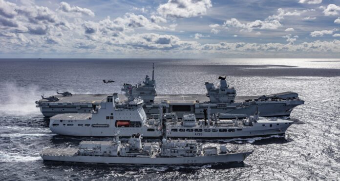 Three Royal Navy vessels, including an aircraft carrier, a support ship, and a frigate, sail closely together on the open sea under a partly cloudy sky.