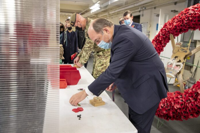A man in a suit and face mask assembles a remembrance poppy at a table, with Army veterans and military personnel standing near a large poppy wreath in the background.