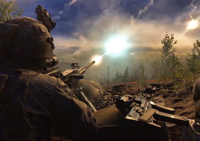 A soldier in Army combat gear fires a weapon at night, muzzle flash and smoke lighting up the dark sky; the area around him includes vegetation, military equipment, and hints of a Pathfinder mission underway.