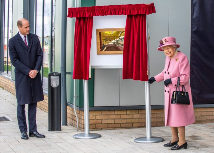 An elderly woman in a pink coat and hat unveils a plaque honoring Army resettlement while a man in a dark coat stands nearby, both outdoors on a paved area next to a building.