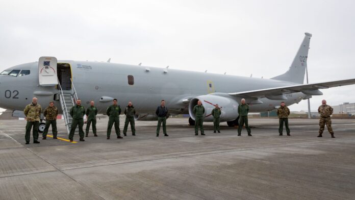 A group of military personnel, including Royal Navy members in green flight suits and others in camouflage uniforms, stands in front of a large gray aircraft on a tarmac.