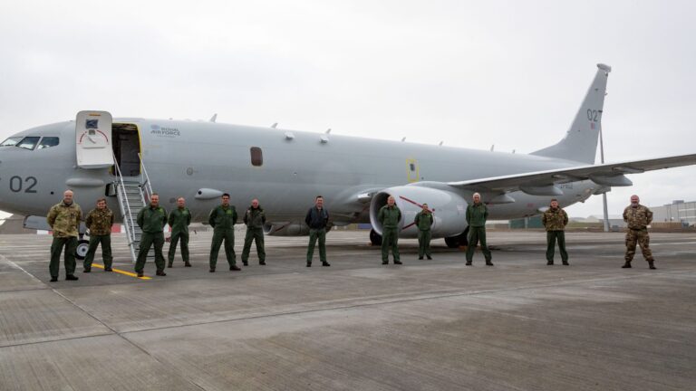 A group of military personnel, including Royal Navy members in green flight suits and others in camouflage uniforms, stands in front of a large gray aircraft on a tarmac.