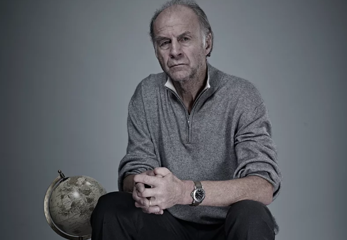 An older Army veteran in a gray zip-up sweater sits with his hands clasped next to a globe, facing the camera against a plain background, reflecting on resettlement and new beginnings.