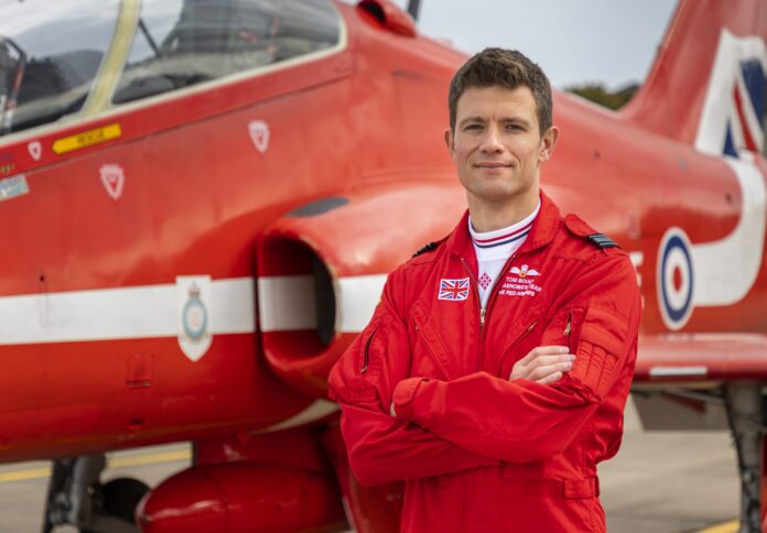 A veteran in a red flight suit stands with arms crossed in front of a red military jet with RAF insignia on the tarmac, embodying the skill and spirit of a true pathfinder.