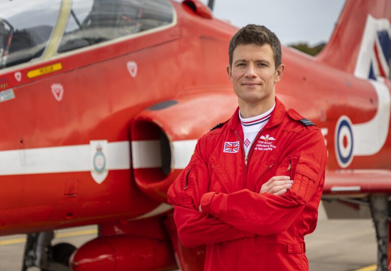 A veteran in a red flight suit stands with arms crossed in front of a red military jet with RAF insignia on the tarmac, embodying the skill and spirit of a true pathfinder.