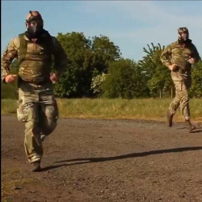 Two people in camouflage uniforms and gas masks, possibly veterans or Pathfinders, running outdoors on a dirt path with grass and trees in the background.