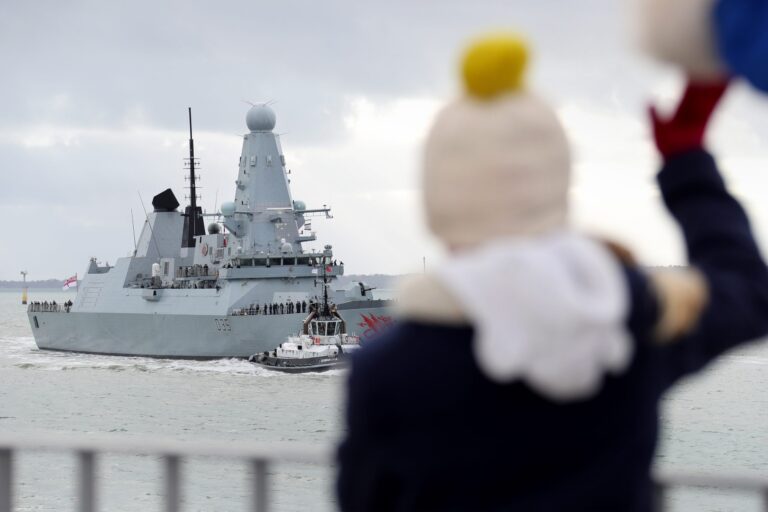 A large Royal Navy ship sails in the water as a person in winter clothing waves from the foreground.