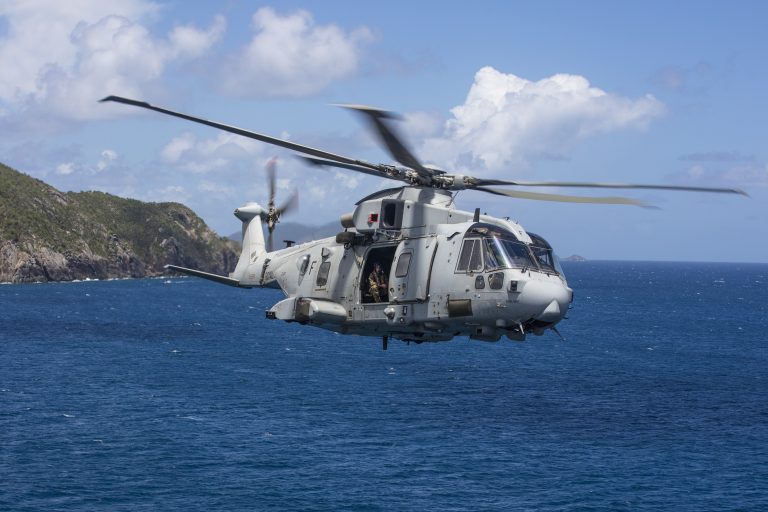 A grey military helicopter flies over the ocean near a coastline under a partly cloudy sky, showcasing the strength and skill of our army veterans.