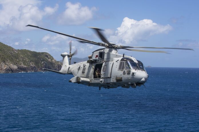 A grey military helicopter flies over the ocean near a coastline under a partly cloudy sky, showcasing the strength and skill of our army veterans.