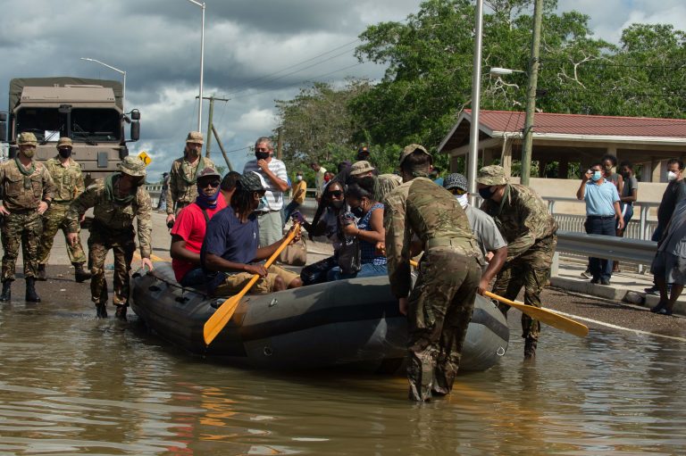 Military personnel assist people in boarding and paddling an inflatable boat on a flooded road, while others observe from the roadside.