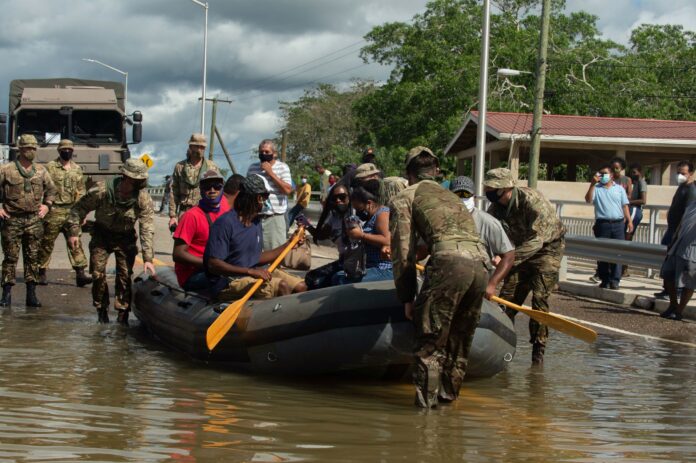Military personnel assist people in boarding and paddling an inflatable boat on a flooded road, while others observe from the roadside.