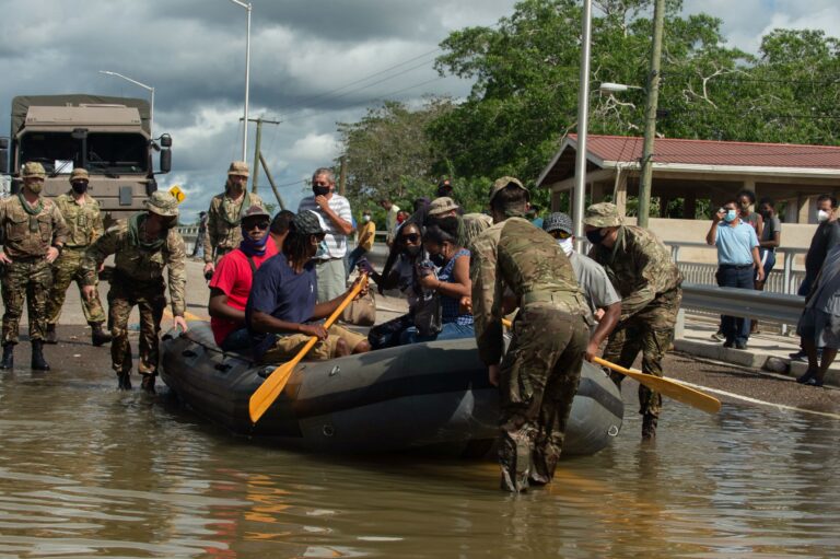 Military personnel assist people in boarding and paddling an inflatable boat on a flooded road, while others observe from the roadside.