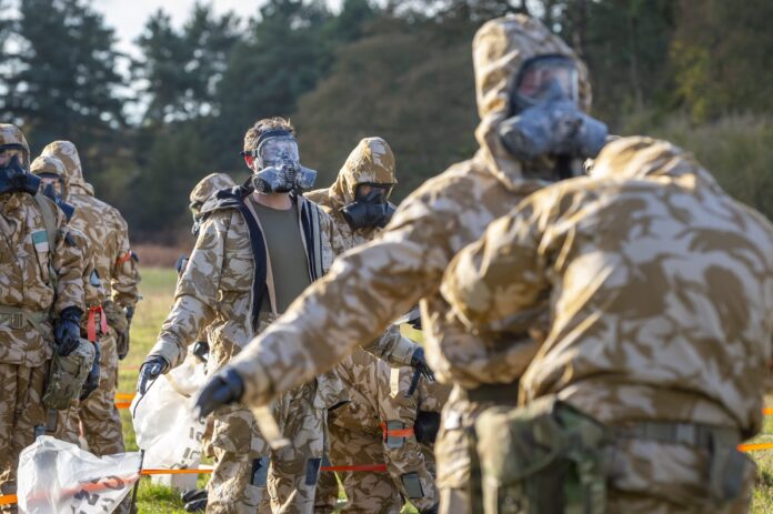 Several people in camouflage hazmat suits and gas masks, possibly Army veterans, stand and interact outdoors, appearing to participate in a decontamination or resettlement safety drill.