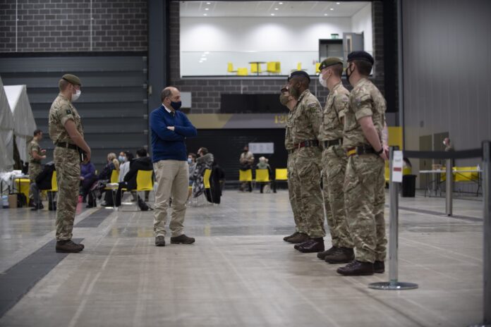 A man in civilian clothes faces four uniformed Military soldiers standing in line inside a large indoor facility with people seated in the background.