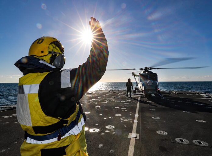 A crew member in safety gear signals a Royal Navy helicopter landing on the deck of a ship at sea under bright sunlight.
