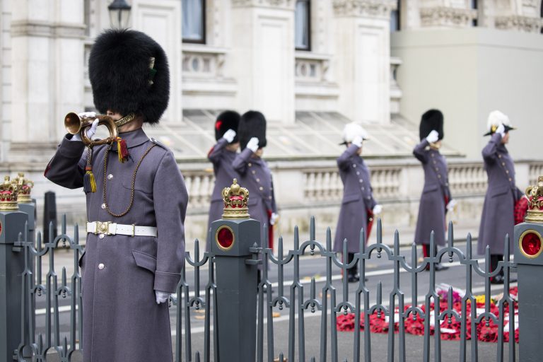 A uniformed military guard plays a bugle while other guards salute near a black fence with gold crowns and red poppy wreaths on the ground; ornate building in the background.