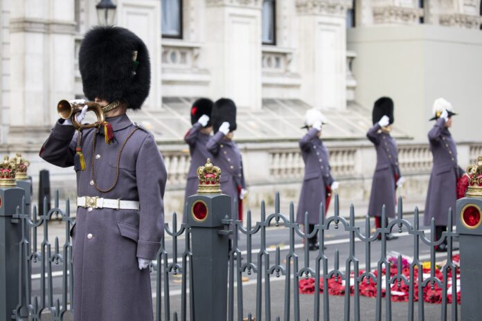 A uniformed military guard plays a bugle while other guards salute near a black fence with gold crowns and red poppy wreaths on the ground; ornate building in the background.