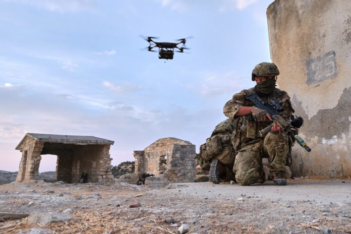 A seasoned Army veteran in camouflage gear kneels behind a wall with a rifle, while a drone hovers in the sky above ruined stone buildings.